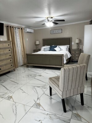 Un dormitorio con cama, sillón, cómoda, mesita de noche, lámpara, ventilador de techo y aire acondicionado instalado en la pared.