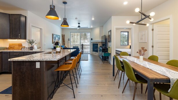 A modern kitchen with dark cabinets, a large island, and a dining area with a wooden table and green chairs. - Luxury Condo, Hot Tub, Balcony, Shuttle to Ski Slopes (Keystone)