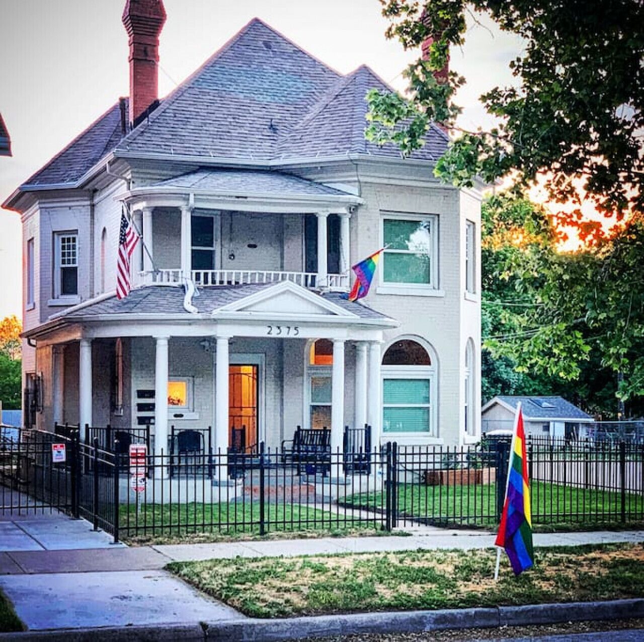 A two-story house with a porch, numbered 2375, featuring a flagpole with an American flag and a rainbow flag.
