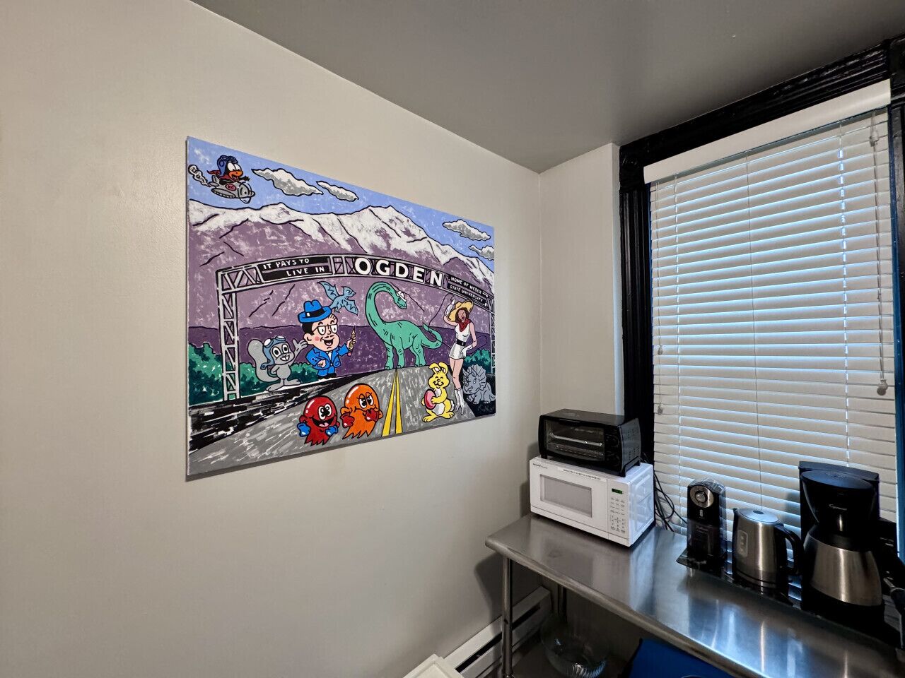 A kitchen with a wall-mounted cartoon mural, a window with blinds, and a coffee maker on the counter.
