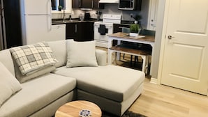 A modern living room with a sectional sofa, a wooden coffee table, and a kitchen area with dark cabinets, a white refrigerator, and a microwave. - Olympic Views - Cozy Modern 2BR Suite (Victoria)