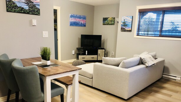 A modern living room with a dining table, grey sofa, and artwork on the walls. - Olympic Views - Cozy Modern 2BR Suite (Victoria)