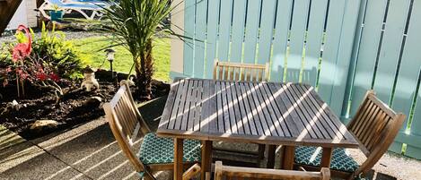 A wooden table with four chairs, a small garden with plants and a statue, and a blue fence in the background.