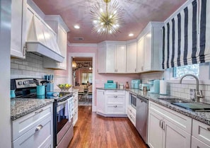 A kitchen with white cabinets, a granite countertop, and a modern chandelier. - Retro FL Charm | Pool + Spa | AMI + Downtown | IMG (Bradenton)