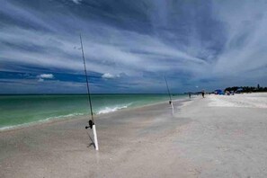 A beach with fishing rods set up on the sand, a clear sky, and the ocean in the background. - Retro FL Charm | Pool + Spa | AMI + Downtown | IMG (Bradenton)