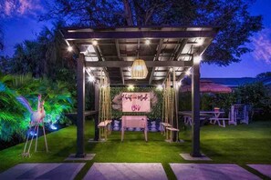 An outdoor gazebo with a table and chairs, surrounded by greenery and a decorative sign.
