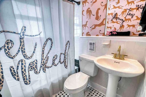 A bathroom with a leopard print shower curtain, a white toilet, a sink with a gold faucet, and a mirror with a leopard print backsplash. - Retro FL Charm | Pool + Spa | AMI + Downtown | IMG (Bradenton)