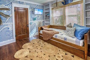 A room with a wooden floor, a bed with a patterned bedspread, a television mounted on the wall, and a wall with a wave pattern. - Retro FL Charm | Pool + Spa | AMI + Downtown | IMG (Bradenton)
