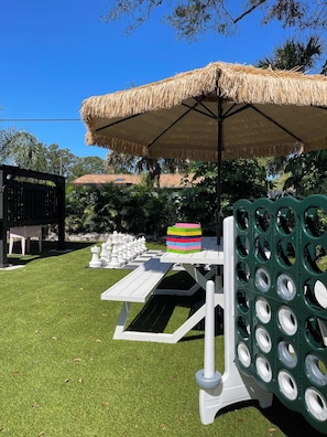 A grassy outdoor area with a thatched umbrella, a chessboard, and a colorful block structure. - Retro FL Charm | Pool + Spa | AMI + Downtown | IMG (Bradenton)