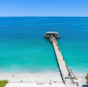 A pier extending into a turquoise sea with a building at the end, a sandy beach, and a clear sky. - Retro FL Charm | Pool + Spa | AMI + Downtown | IMG (Bradenton)