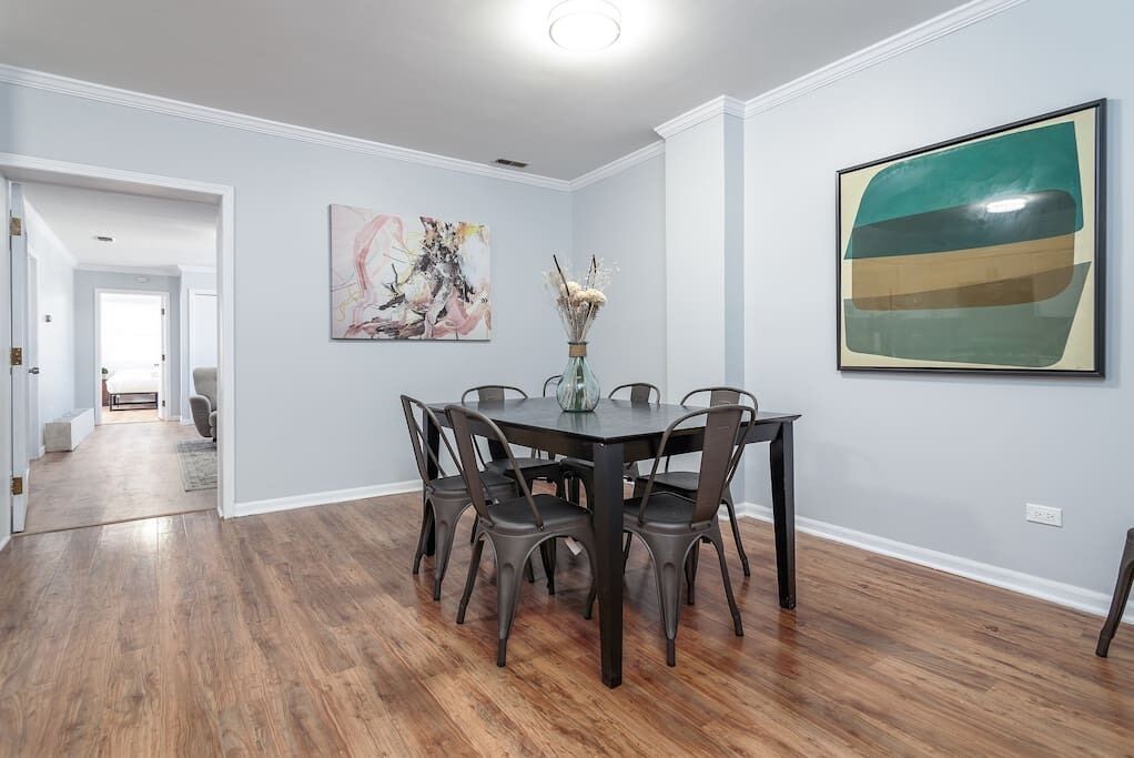 A dining area with a dark wooden table and chairs, a vase with dried flowers, and two abstract paintings on the walls.