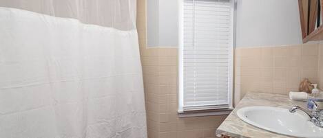 A bathroom with a shower curtain, a window with blinds, a sink, and a mirror.