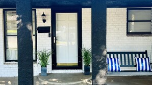 Entrada de un edificio con una puerta blanca, ventanas de marco negro y un banco con cojines a rayas.