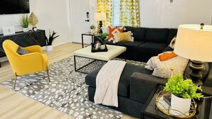 Una sala de estar moderna con un sofá negro, un sillón amarillo, una mesa de centro y un televisor montado en la pared.