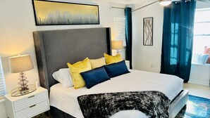 Un dormitorio con cama, mesitas de noche, lámpara, un cuadro y ventana con cortinas.