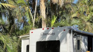 Un camping-car blanc, portant l’inscription « Cherokee » à l’arrière, est garé dans un jardin luxuriant, agrémenté de palmiers et de diverses plantes.