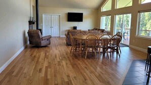 Un vaste salon doté d’un plancher en bois, d’une cheminée, d’un coin repas avec une table et des chaises, et de grandes fenêtres offrant une vue sur l’extérieur.