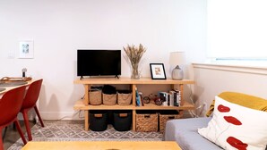 Un salon avec un téléviseur posé sur une étagère en bois, un vase contenant des plantes séchées et une bibliothèque remplie de livres et de paniers.