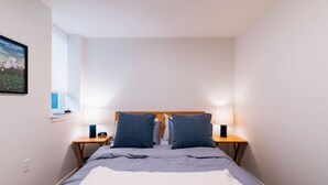Une chambre à coucher avec un lit, deux tables de chevet avec des lampes, une fenêtre avec des stores et un tableau accroché au mur.