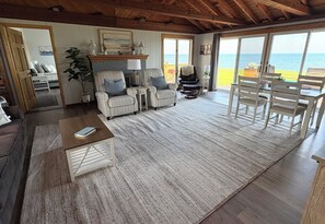 A spacious living area with a large window overlooking the sea, a fireplace, striped armchairs, and a dining set.