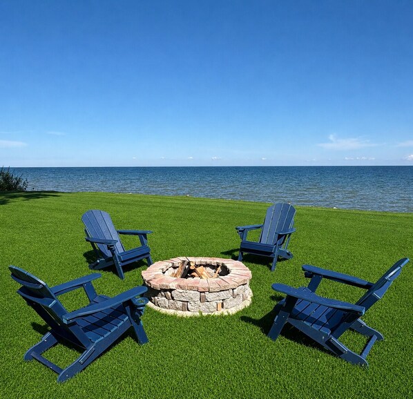 A fire pit surrounded by four Adirondack chairs on a grassy lawn with a view of the ocean.
