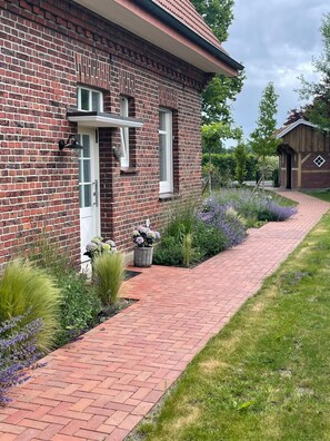 A brick house with a red brick pathway leading to the entrance, surrounded by well-maintained gardens and a small wooden structure. - Quiet vacation home in a rural setting with a garden in the Münsterland region (Gescher)