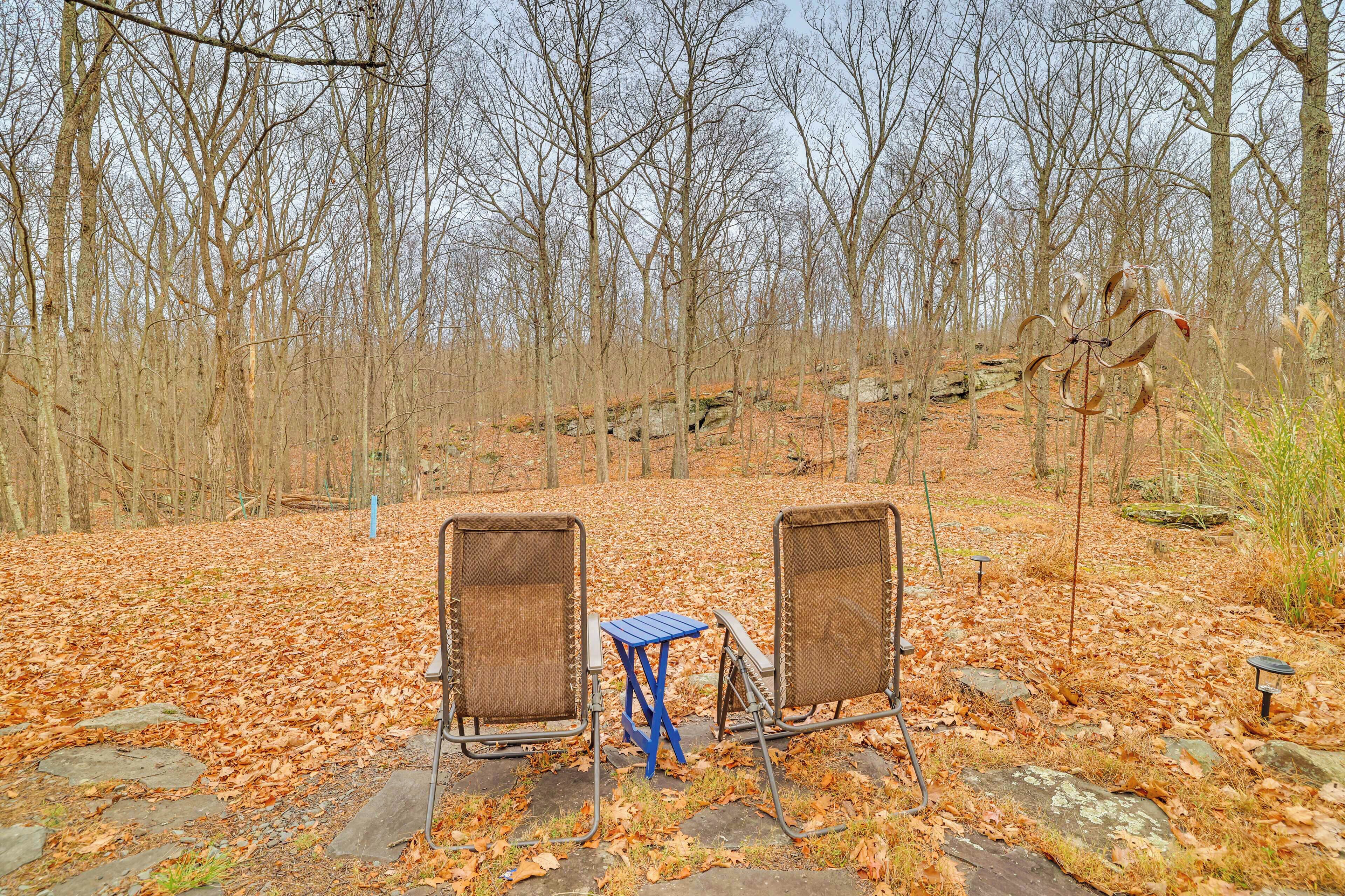 Two wicker chairs and a small blue table set on a stone path in a forested area with leaf-covered ground.