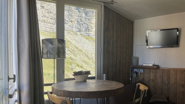 A cozy room with a round wooden table, two chairs, a TV mounted on the wall, and a window with a view of a rocky hillside. - Apartment at the foot of the slopes (Morzine)