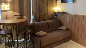 A cozy room with a wooden floor, a brown sofa with cushions, a round wooden table with chairs, a lamp, and a TV mounted on the wall. - Apartment at the foot of the slopes (Morzine)