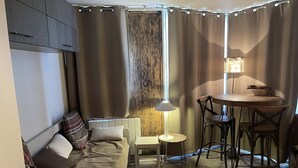 A compact living space with a sofa, a small table with chairs, a lamp, and a window with curtains. - Apartment at the foot of the slopes (Morzine)