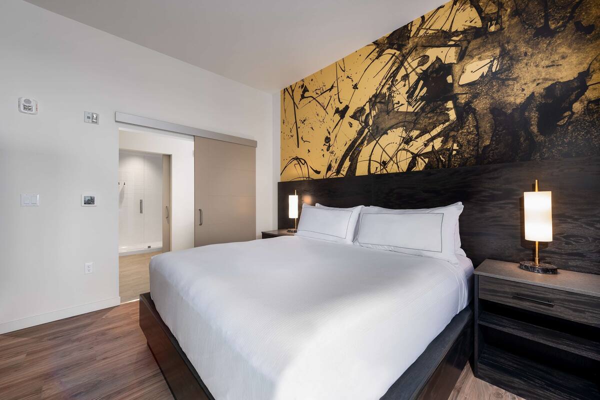 Ein modernes Hotelzimmer mit einem großen Bett, einem dunklen Kopfteil und einer Wand mit abstrakter Kunst.