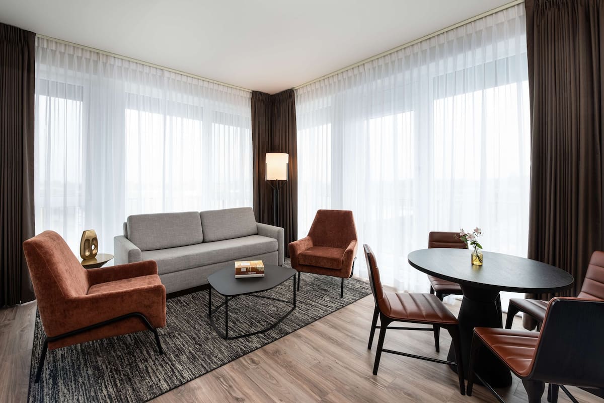 Ein modernes Hotelzimmer mit grauem Sofa, zwei Sesseln, einem runden Esstisch und einer Stehlampe.