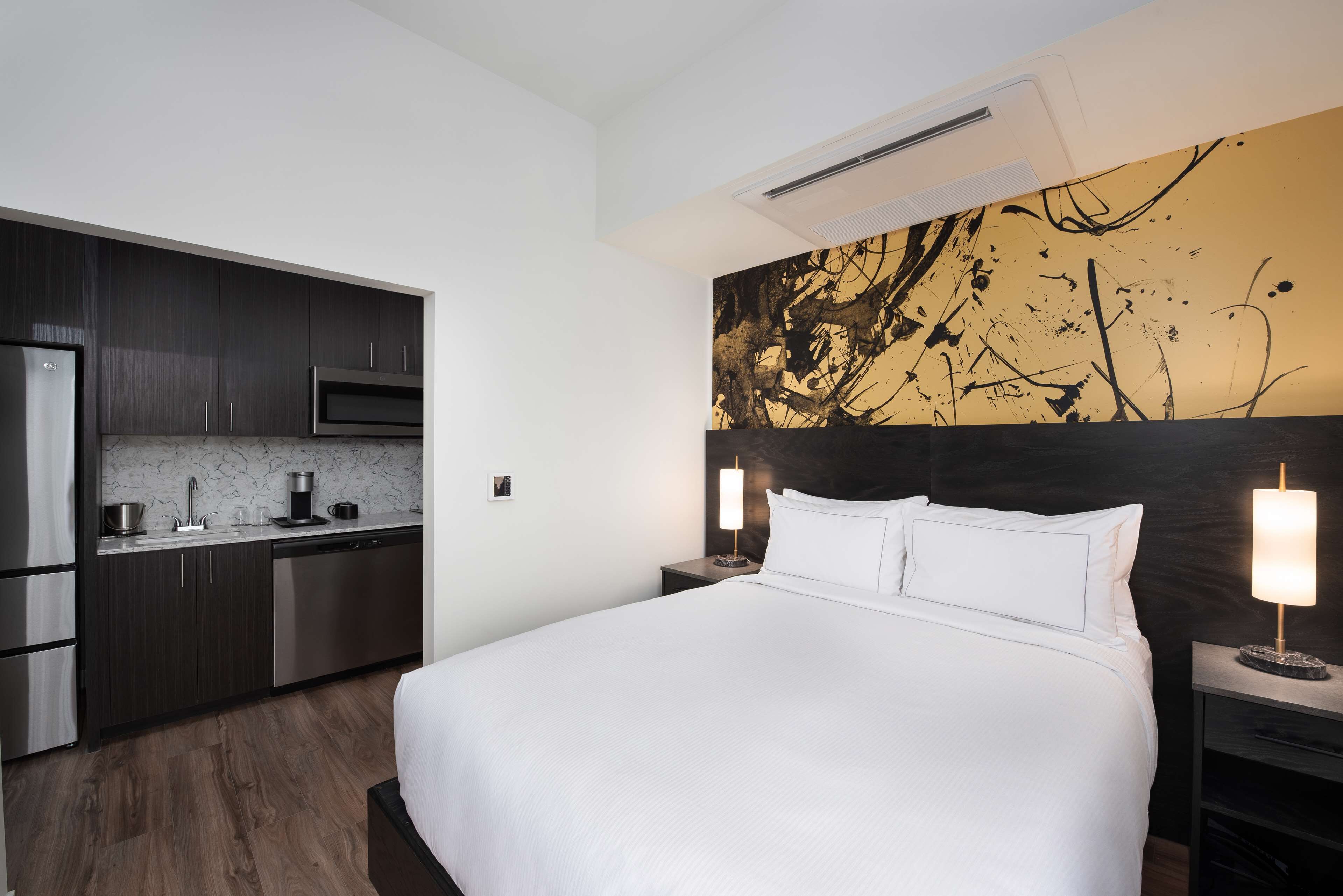 Una habitación de hotel moderna con una cama grande, una kitchenette y una original obra de arte abstracta en la pared.