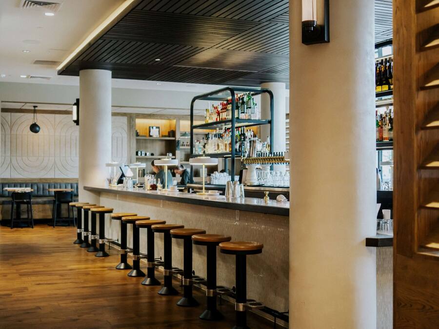 A bar area with wooden stools, a counter with bar tools, and shelves with bottles.