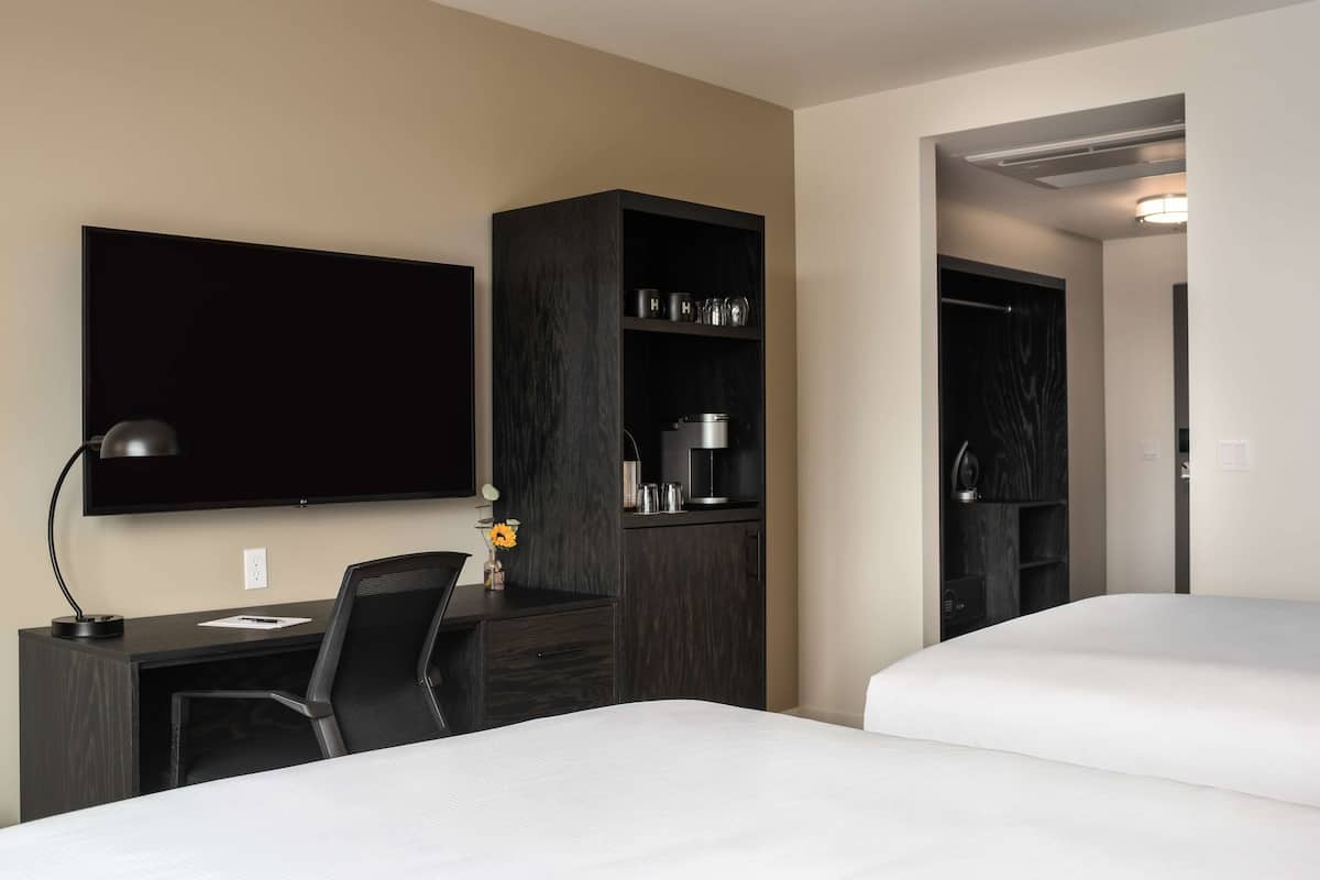 Ein Hotelzimmer mit einem Bett, einem Schreibtisch mit Stuhl, einem an der Wand montierten Fernseher, einem Kleiderschrank und einem Regal mit Küchenutensilien.