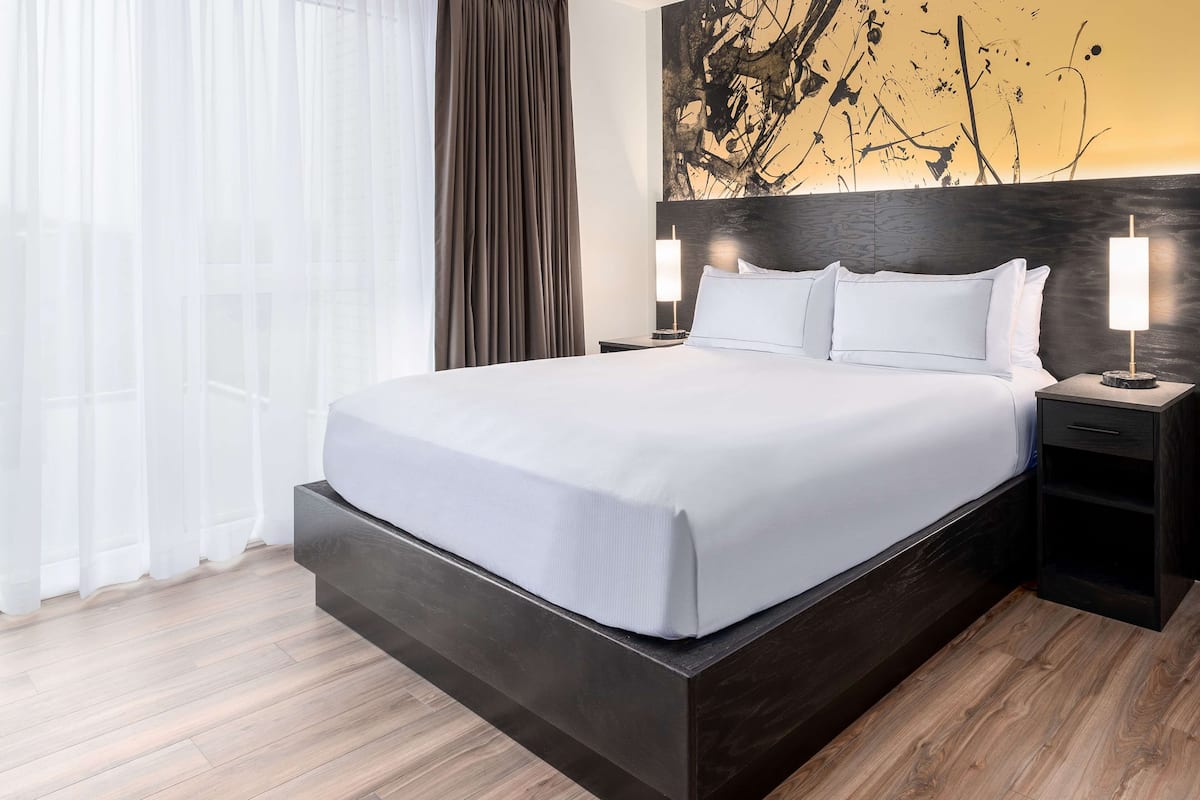 Ein modernes Hotelzimmer mit einem großen Bett, einem dunklen Holz-Kopfteil, weißen Bettwäsche und einem Bild an der Wand.