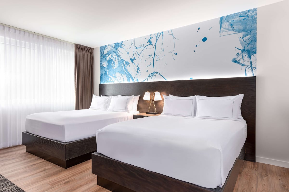 Ein Hotelzimmer mit zwei Betten, einem großen abstrakten Gemälde an der Wand und einem Fenster mit transparenten Vorhängen.