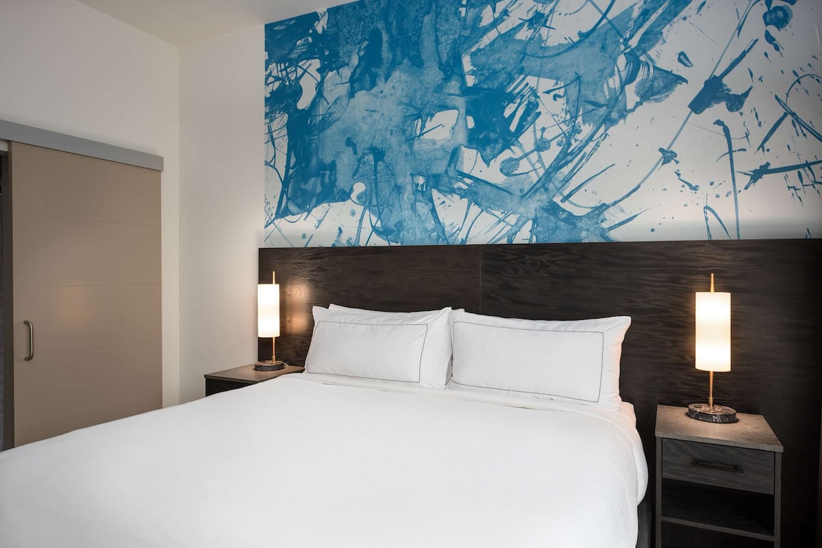 Ein Hotelzimmer mit einem großen Bett, zwei Nachttischen mit Lampen, einem dunklen Kopfteil und einem großen, abstrakten blauen Gemälde an der Wand.