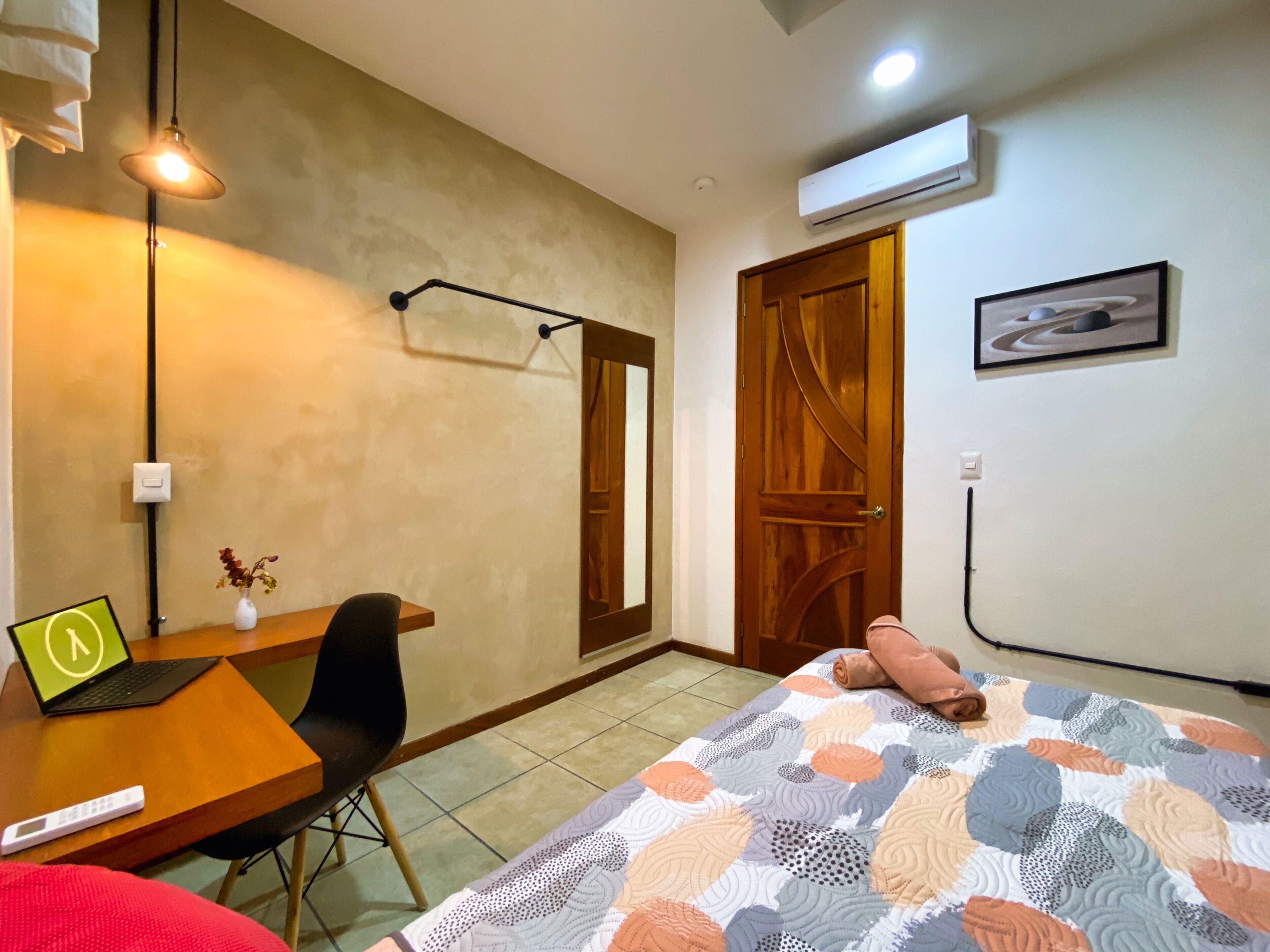 A bedroom with a bed, a desk with a laptop, a chair, a bedside table, a wall-mounted air conditioner, and a door.