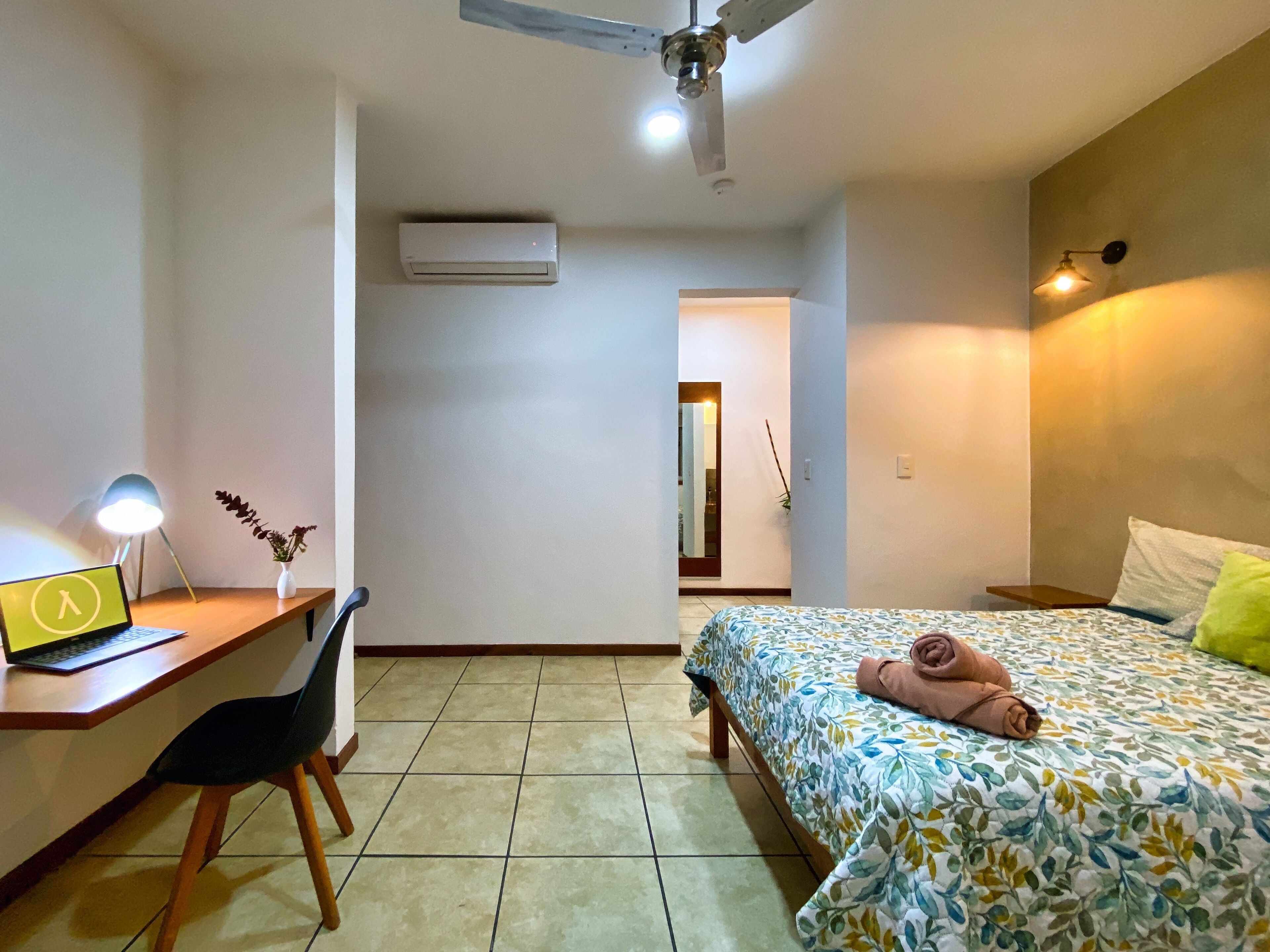 Quarto com cama, escrivaninha com laptop, cadeira, ar condicionado e ventilador de teto.