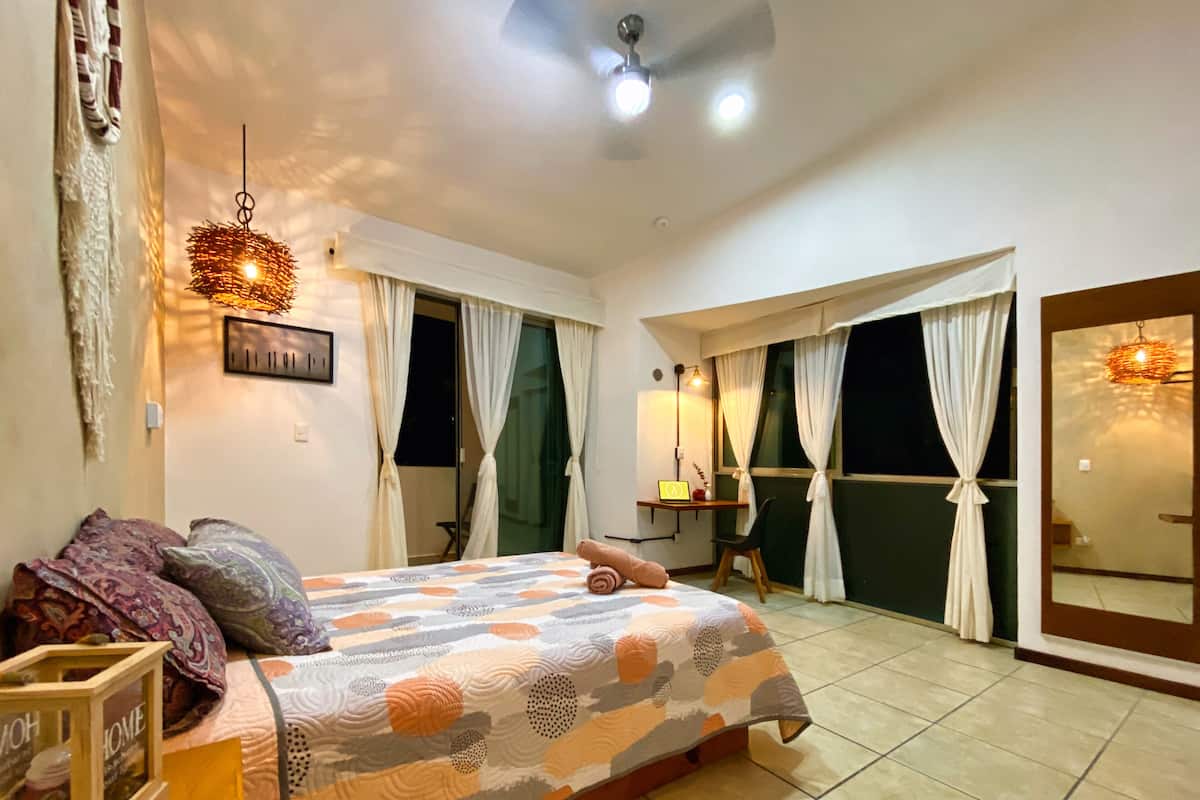 A bedroom with a bed, a desk, and a chair. There is a window with curtains, a ceiling fan, and a wall-mounted light fixture.