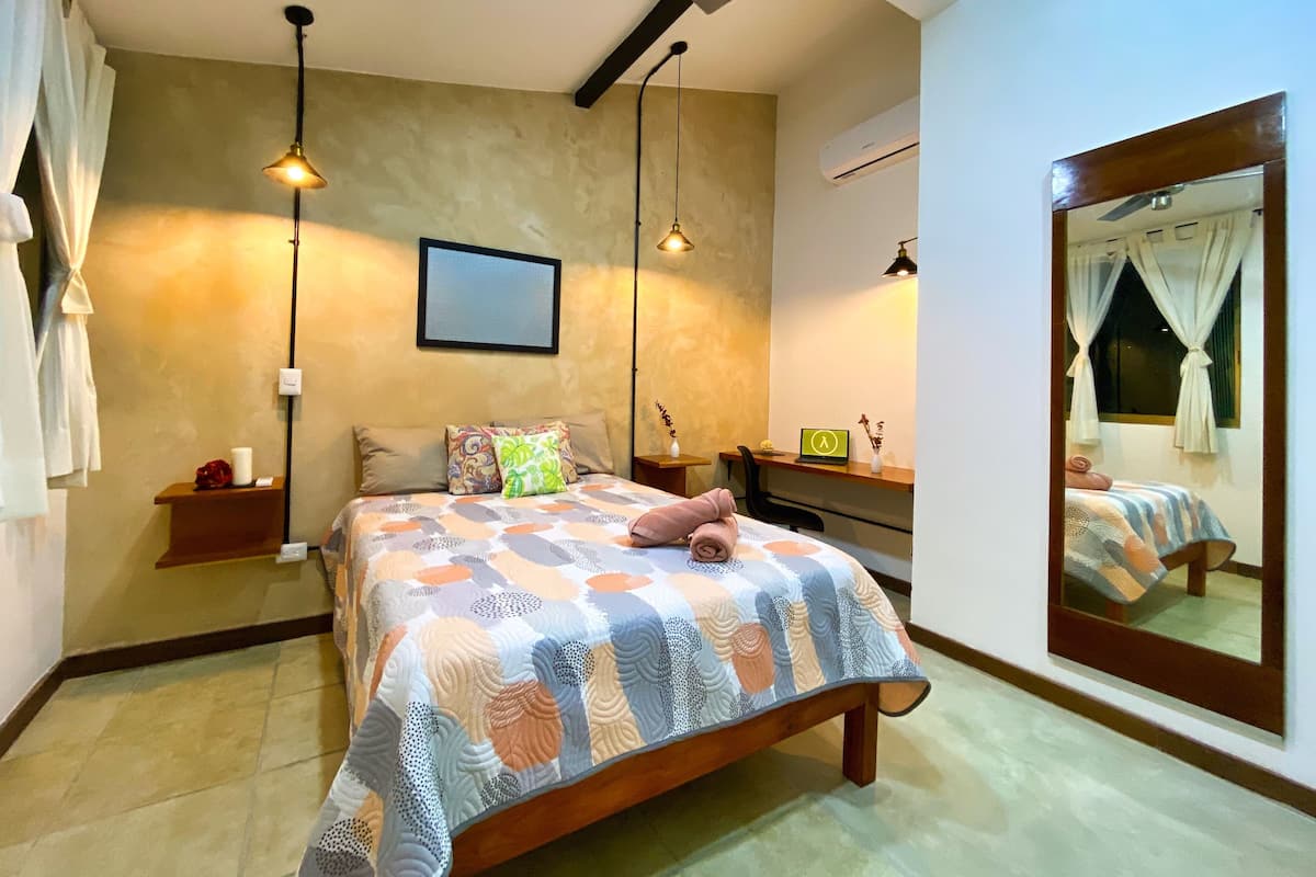 A bedroom with a bed, bedside tables, a desk, a chair, a mirror, and a ceiling light fixture.