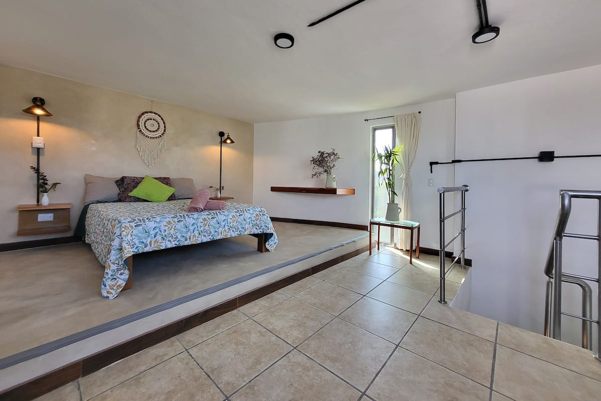 A bedroom with a bed, bedside lamps, a wall-mounted shelf, a potted plant, and a ladder.