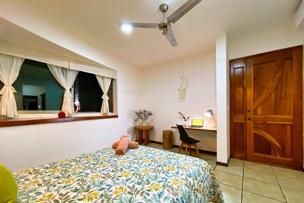 A bedroom with a bed, a desk, a chair, a ceiling fan, and a wooden door.