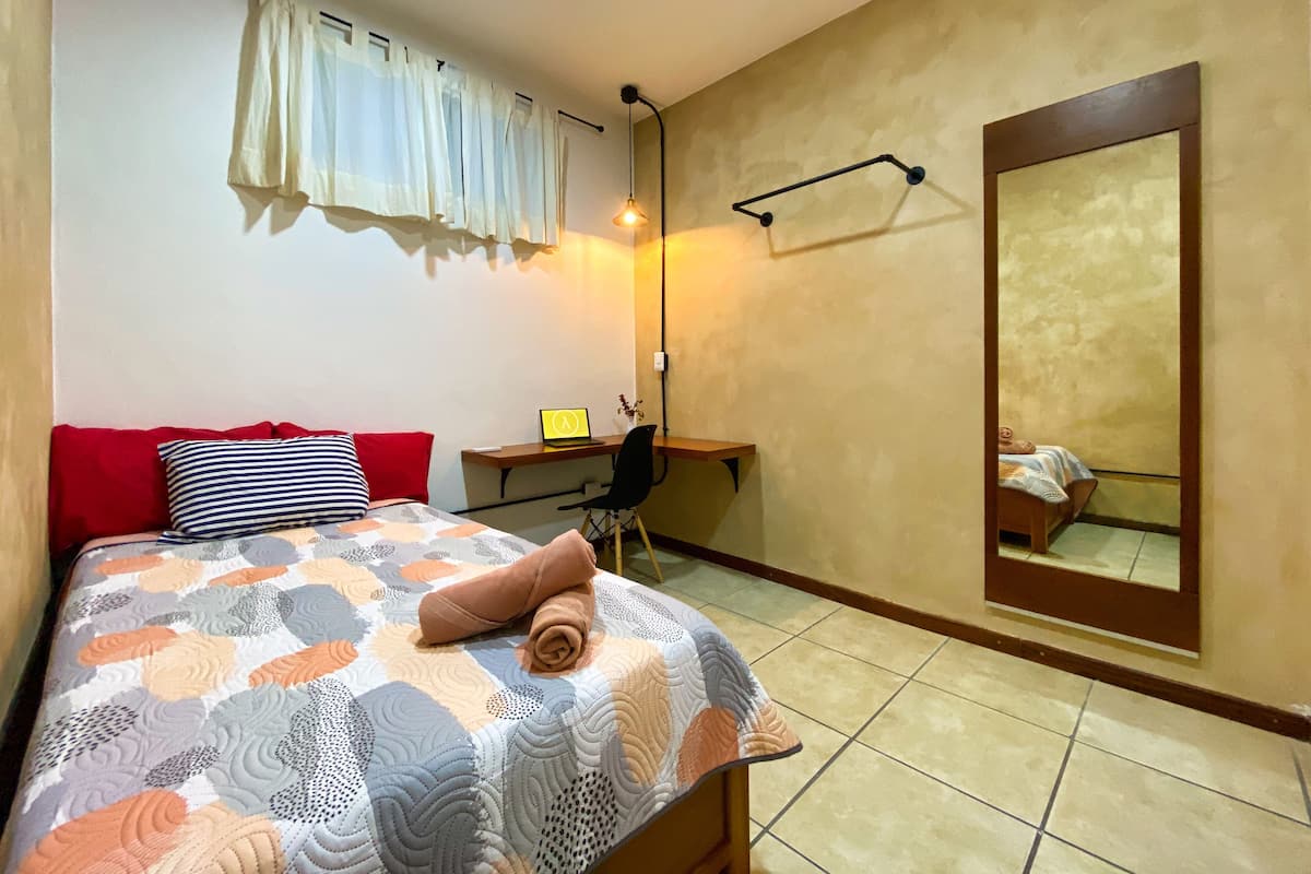 A bedroom with a bed, a desk, a chair, a lamp, a mirror, and a window with curtains.