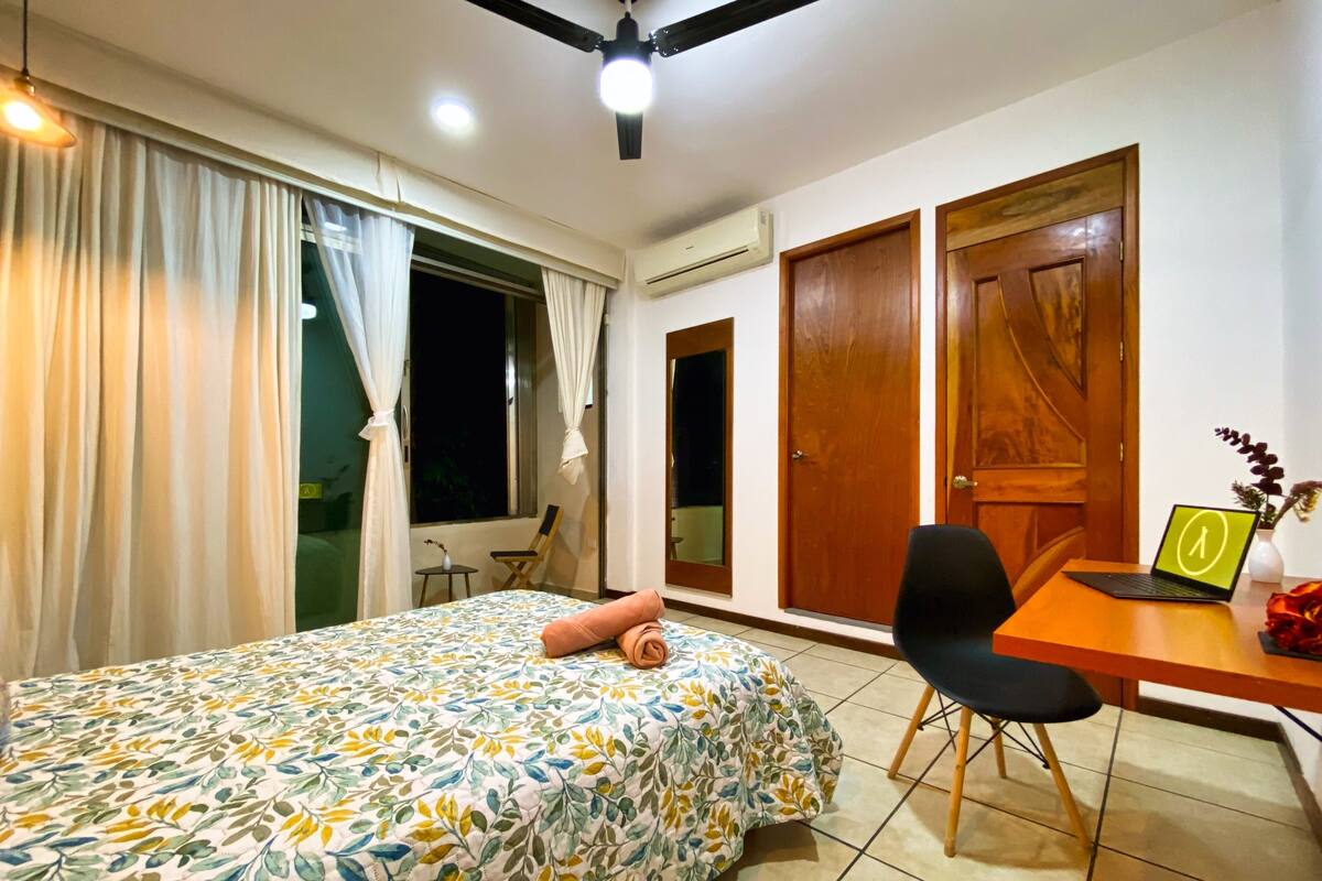 A bedroom with a bed, a desk, a chair, and a window with curtains.