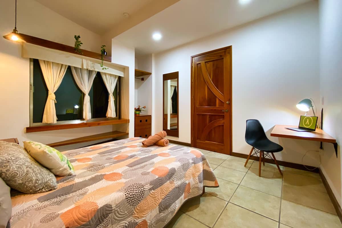 A bedroom with a bed, a desk, a chair, a mirror, and a door.