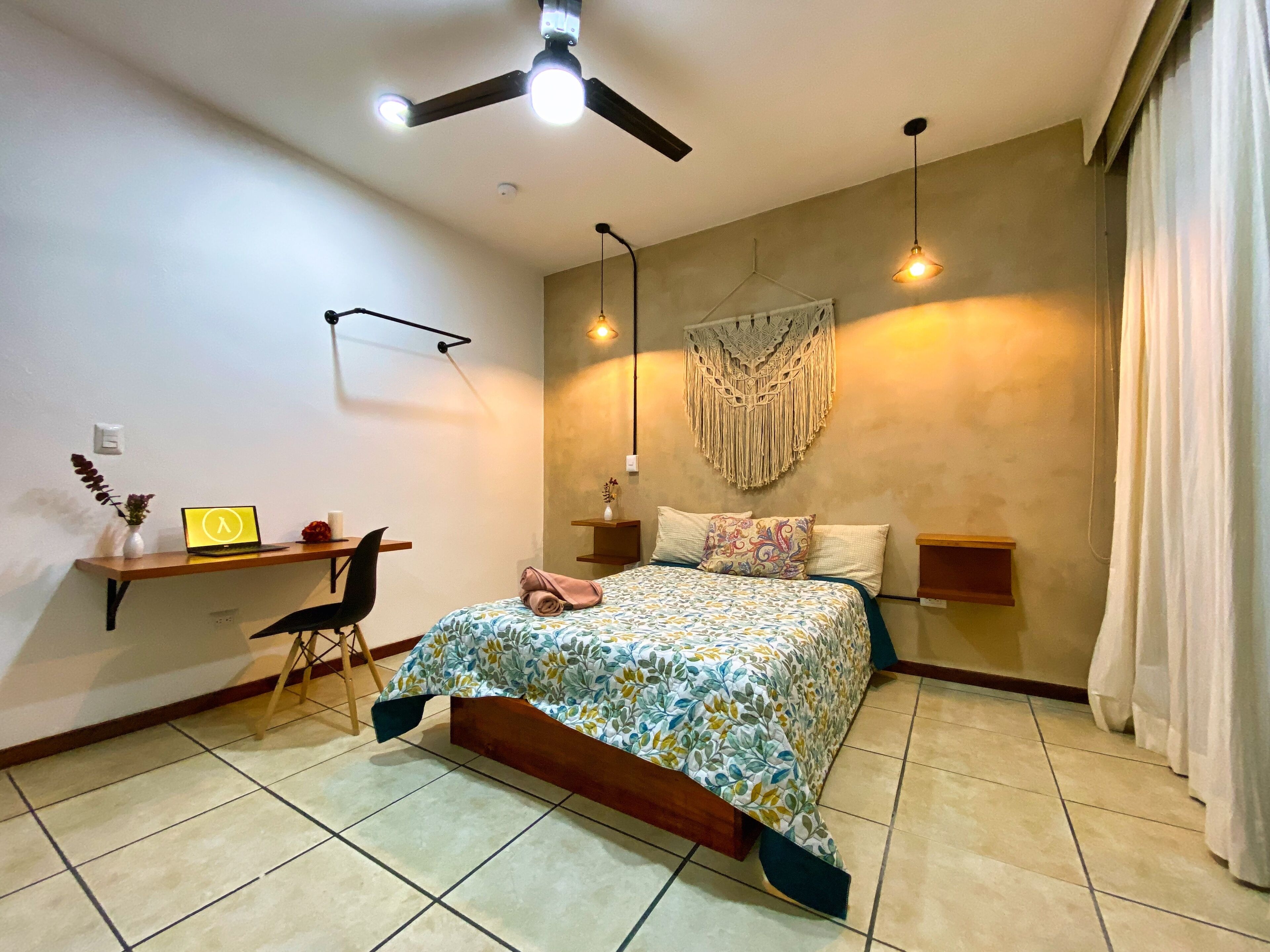 Un dormitorio con cama, mesitas de noche, un escritorio con una computadora portátil, una silla, un ventilador de techo y un adorno de macramé en la pared.
