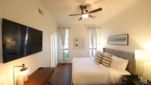 Quarto de hotel com cama, televisão, ventoinha no teto e janela com cortinas.