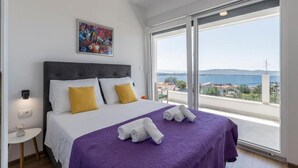 A bedroom with a bed, a nightstand, a painting, and a large window with a view of the sea and buildings. - Luxury Villa La nonna Ana - entertainment,fitness,pool,sea view (Kastel Sucurac)